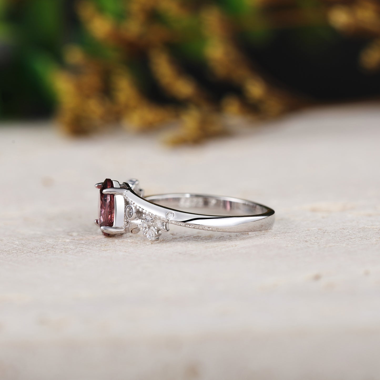 White gold ring with a red gemstone on a light surface with blurred greenery in the background
