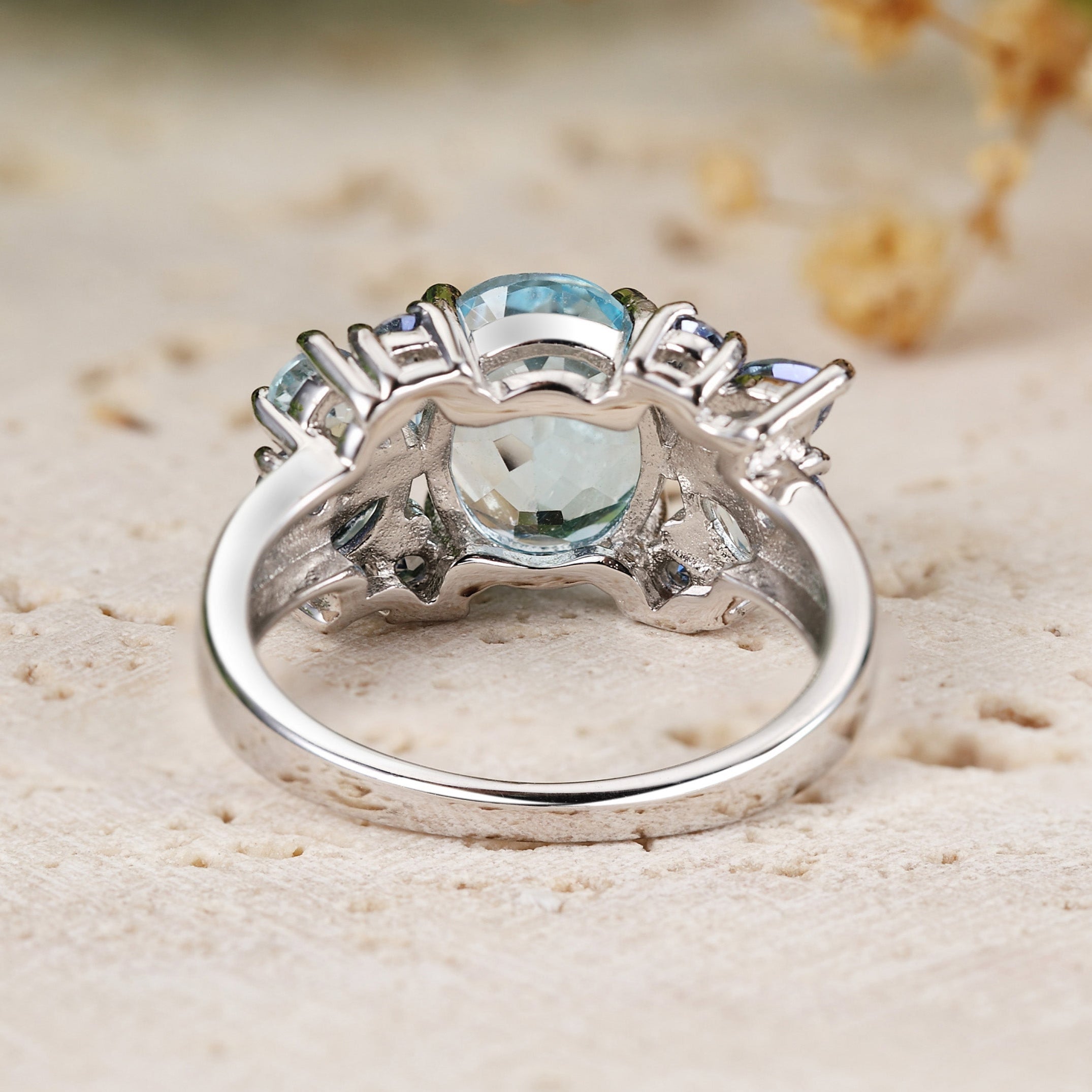 White gold ring with a central gemstone on a sandy surface with blurred flowers in the background