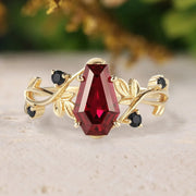 Gold ring with a red gemstone and black stones on a beige surface with blurred greenery in the background